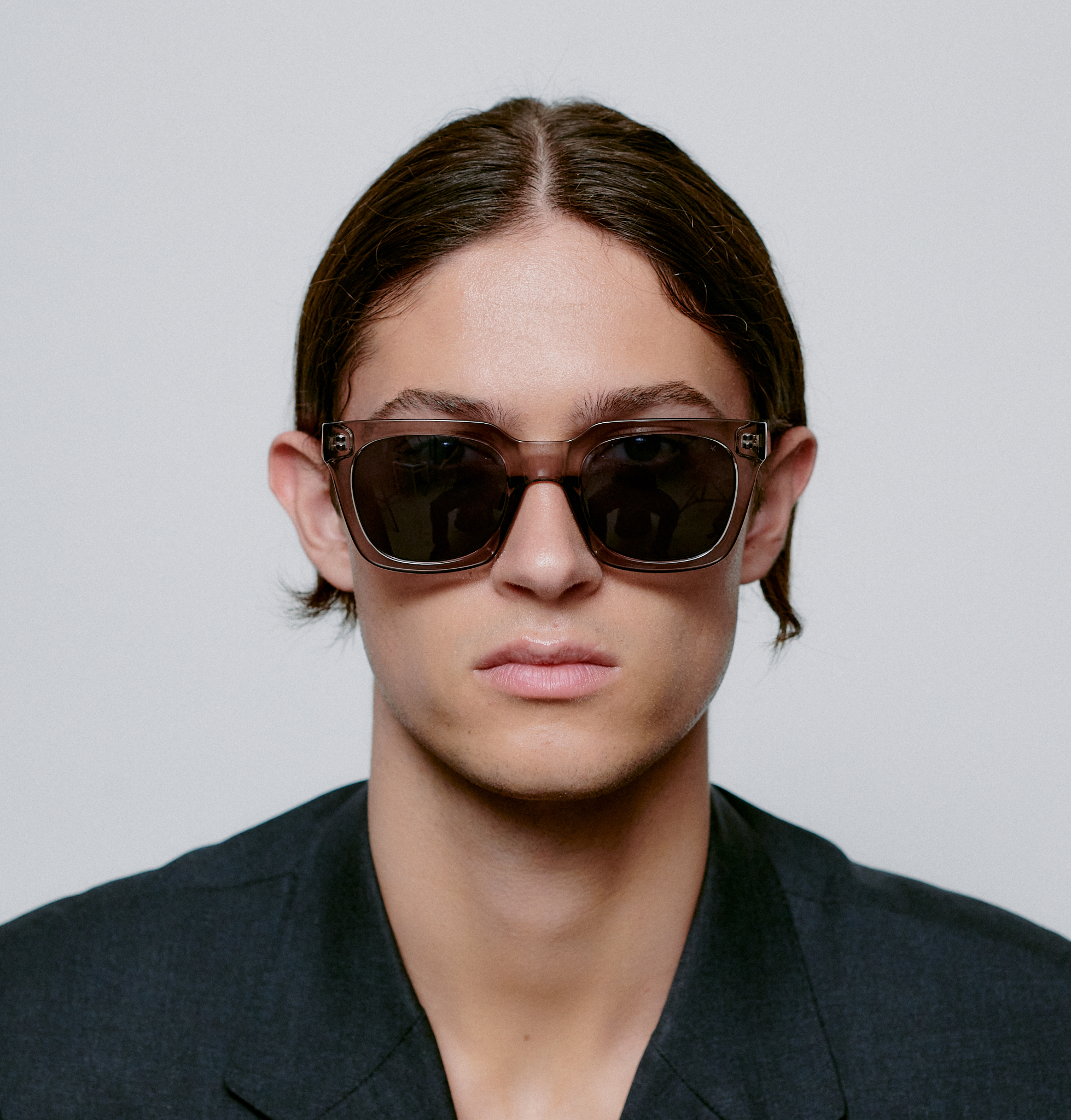 A person with straight brown hair parted in the middle wears NANCY - Grey Transparent sunglasses and a dark blazer, embodying Estonian design as they look directly at the camera against a plain light background. Gate21.ee