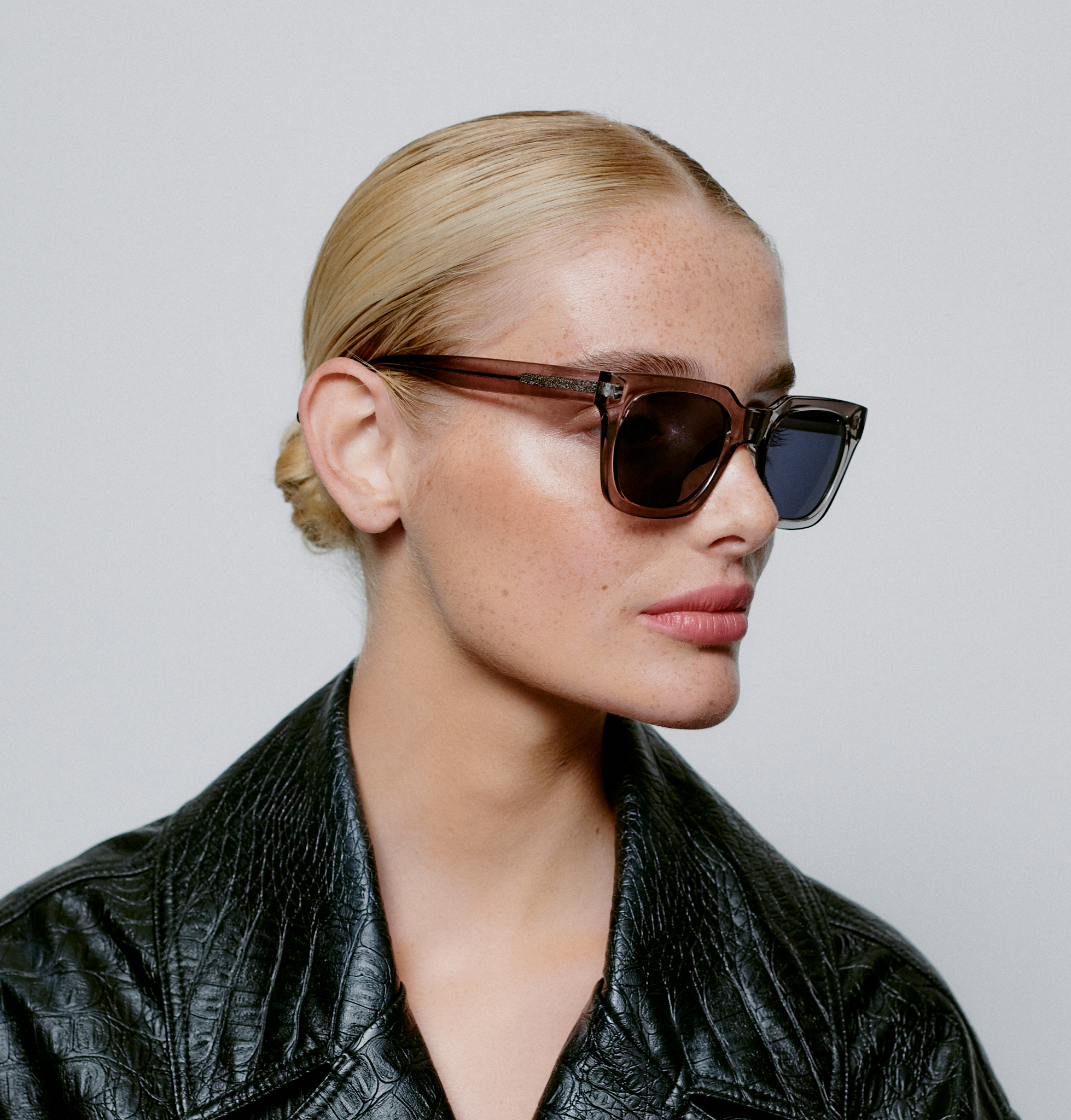 A person with light skin and blonde hair in a low bun wears the NANCY - Grey Transparent sunglasses and a black textured jacket, highlighting curated Estonian design. The plain, light background features the person facing slightly to the right. Gate21.ee