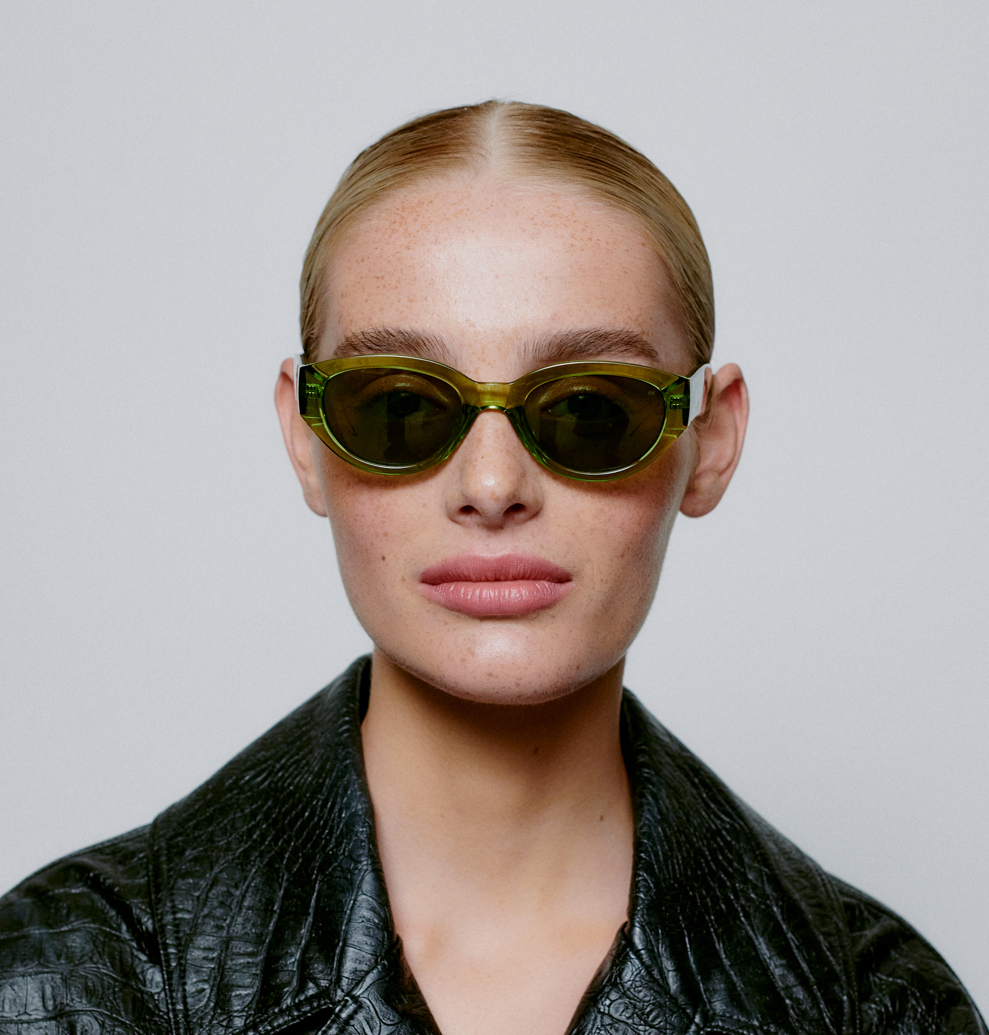 A person with slicked-back blonde hair wears WINNIE - Light Olive Transparent sunglasses and a black textured jacket, looking at the camera against a plain light grey background—an image echoing sleek Estonian design. Gate21.ee