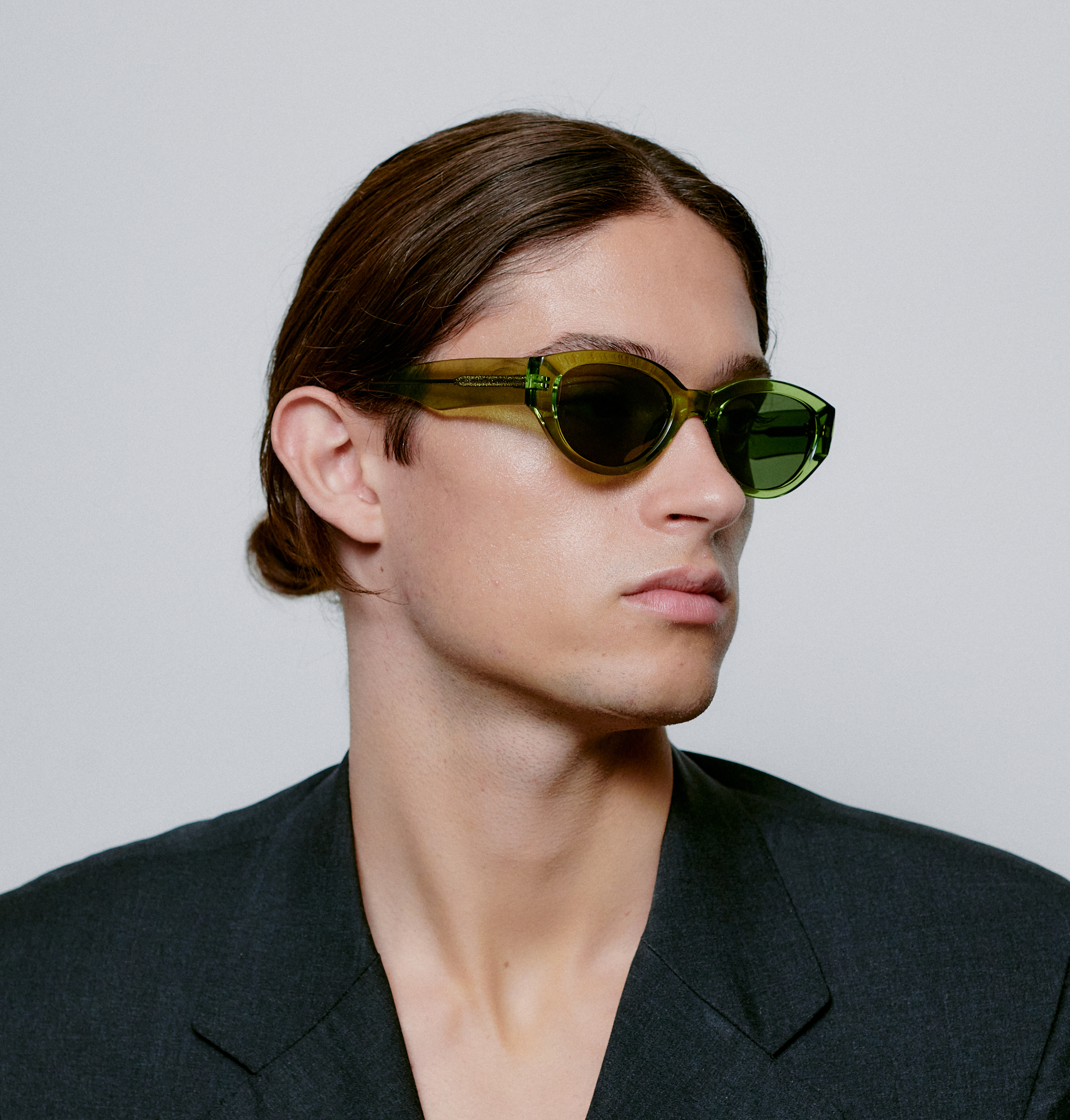 A person with straight brown hair pulled back, wearing WINNIE - Light Olive Transparent oval sunglasses and a dark blazer, looks to the left against a plain light background—embodying curated Estonian design and unique style. Gate21.ee