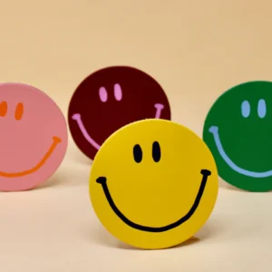 Four round Leather Coaster - Ark Design - KÕIK smiley face cut-outs in pink, maroon, green, and yellow are displayed on a light beige background. The Estonian-designed yellow smiley is in front, with the others slightly behind. Gate21.ee