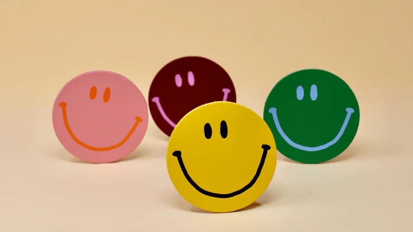 Four round Leather Coaster - Ark Design - KÕIK smiley face cut-outs in pink, maroon, green, and yellow are displayed on a light beige background. The Estonian-designed yellow smiley is in front, with the others slightly behind. Gate21.ee
