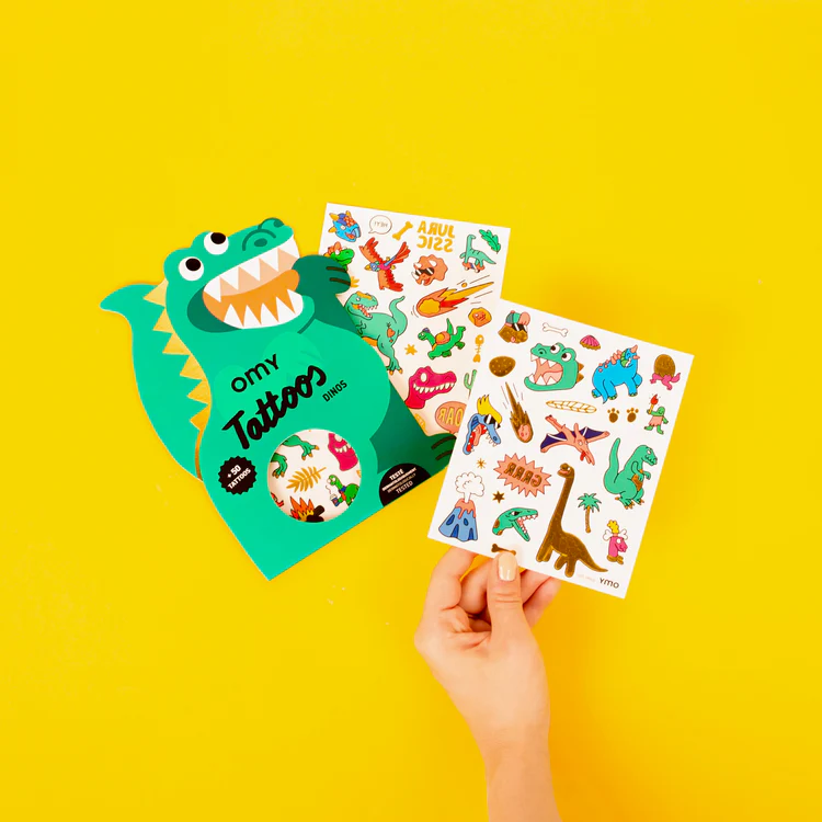 A hand displays two vibrant Dinos – Temporary Tattoo sheets beside a green dragon-shaped package on a bright yellow background—great for gift shops or Tallinn shopping adventures. Gate21.ee