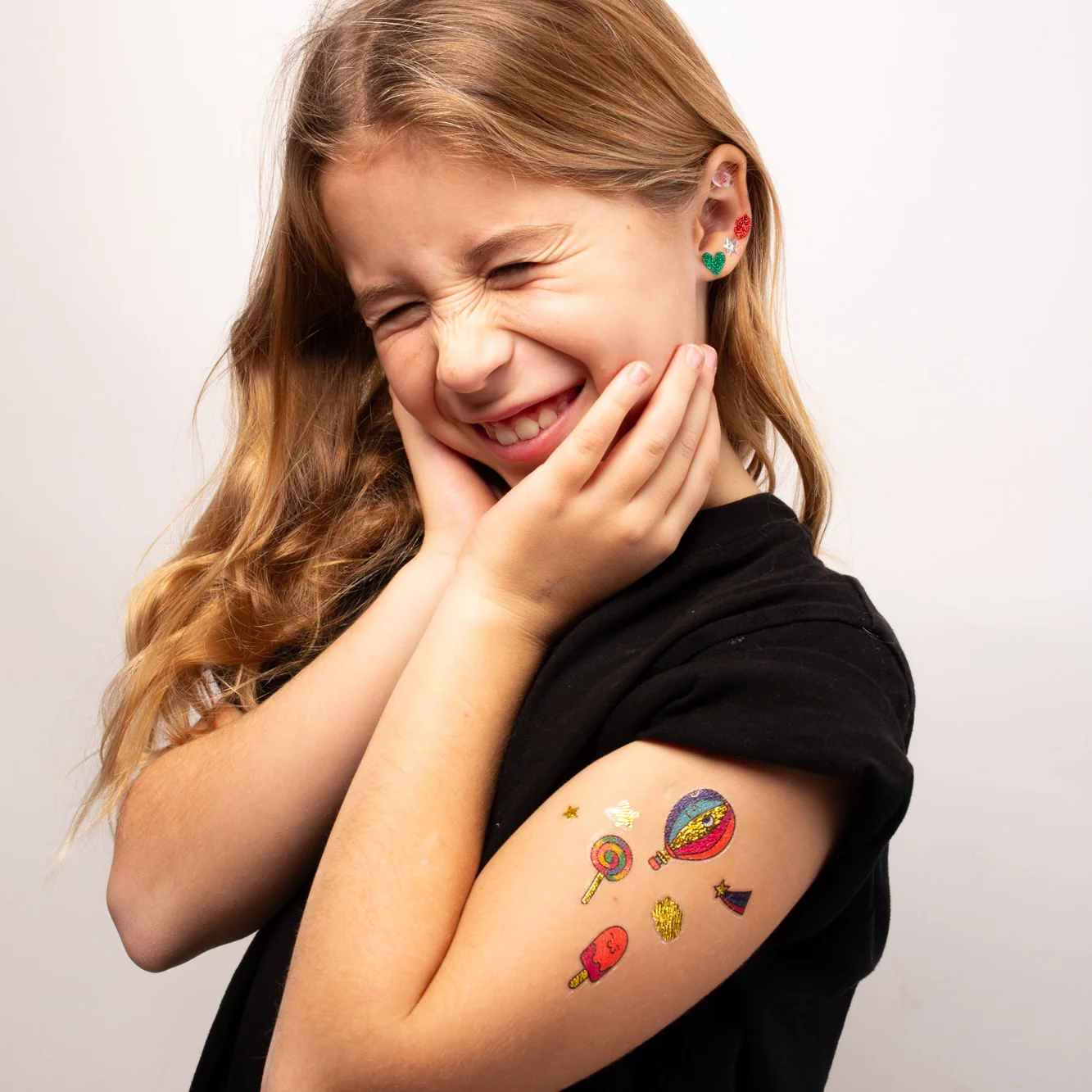 A smiling young girl with long blonde hair, eyes closed and hands on cheeks, wears a black shirt and showcases Kawaii - Temporary Tattoos featuring planets, rockets, lollies, ice lollies, and stars in playful Estonian design. Gate21.ee