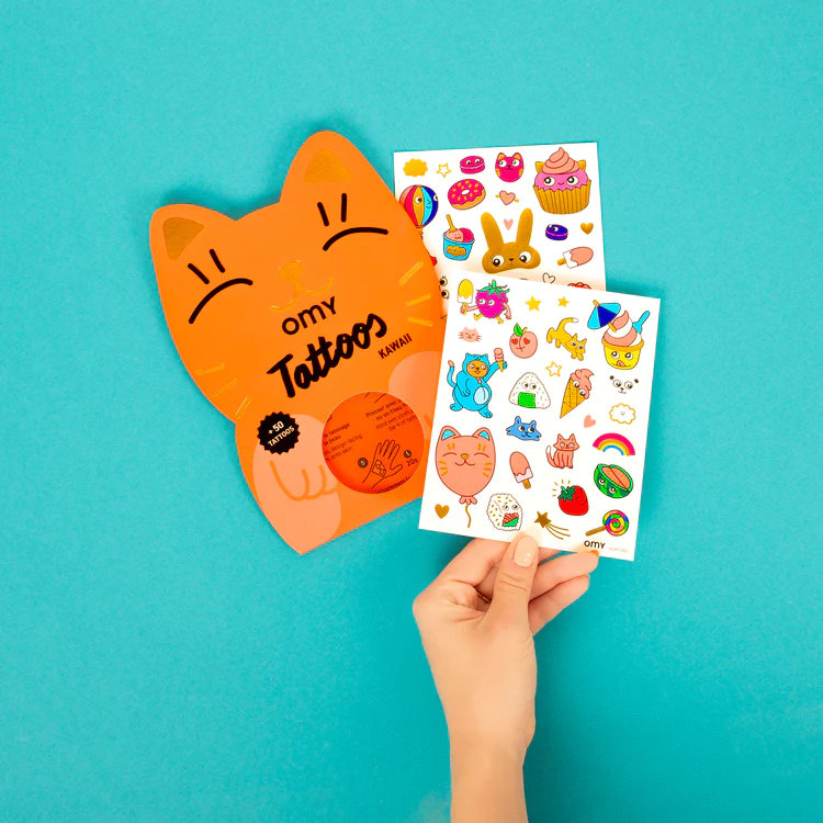 A hand displays two sheets of Kawaii - Temporary Tattoos with playful cartoon designs, beside vibrant orange cat-shaped packaging marked "omy Tattoos"—a quirky Tallinn gift idea against a turquoise background. Gate21.ee