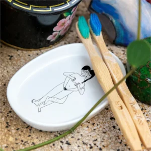 Two bamboo toothbrushes with blue and green bristles sit on the Faces - Tray, which features an illustrated person. The tray is on a speckled surface next to a floral black container and a blue object—ideal for a curated gift shop display. Gate21.ee