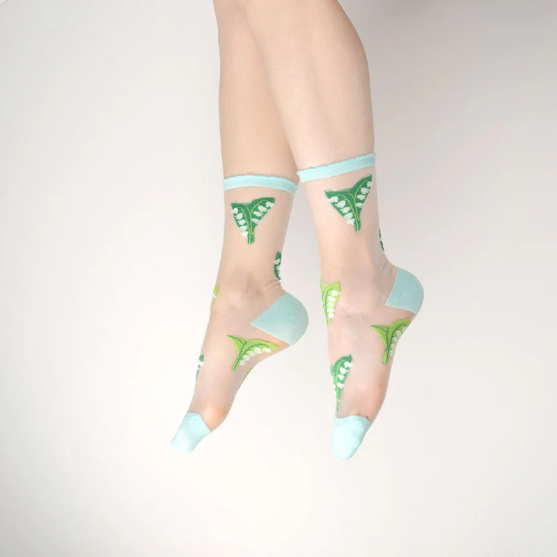 A pair of feet in Lily of the Valley - Transparent Socks, featuring green butterfly designs with light blue heels, toes, and cuffs—ideal for curated outfits or style inspiration whilst shopping in Tallinn, set against a simple light background. Gate21.ee