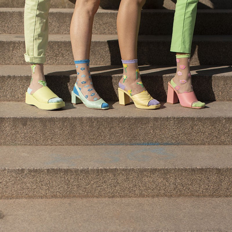 Three people stand on stone steps, each wearing pastel open-toe heels and Lily of the Valley - Transparent Socks—a playful display of curated Estonian-inspired style. Only their legs and feet are visible. Gate21.ee