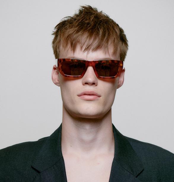 Wearing the FAME - Havana large rectangular sunglasses and a dark blazer, a person with short, tousled light brown hair channels Estonian-inspired style against a plain light background. Gate21.ee