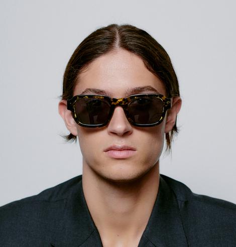 A person with straight brown hair, parted in the middle, wears HALO - Demi tortoiseshell sunglasses and a black blazer, capturing modern Estonian style as they face forwards against a plain light background. Gate21.ee