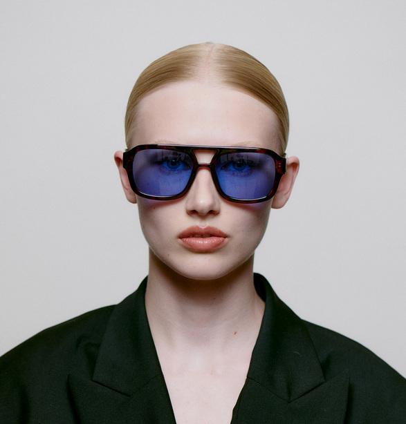 A light-skinned person with blonde hair pulled back wears a dark blazer and KAYA - Demi tortoiseshell blue lenses sunglasses, facing the camera against a plain background—embodying modern style perfect for a chic gift shop. Gate21.ee