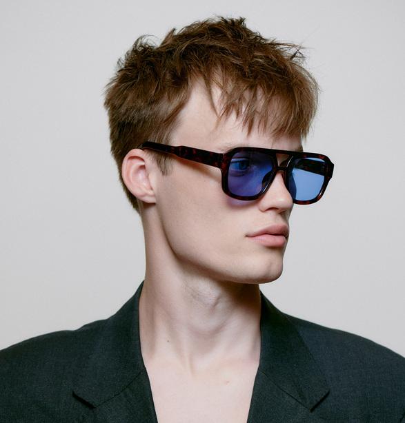A young person with short, tousled brown hair wears KAYA - Demi tortoiseshell blue lenses sunglasses and a dark blazer, gazing to the side against a plain light background, capturing the essence of modern Estonian design from Tallinn shopping districts. Gate21.ee