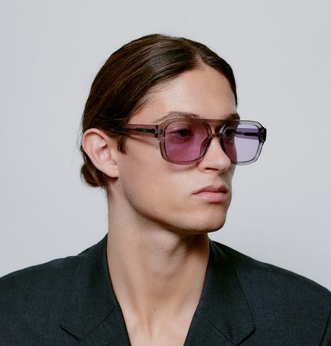 A person with long brown hair tied back wears the KAYA - Grey Transparent oversized square sunglasses, featuring grey transparent frames and purple-tinted lenses, paired with a dark blazer—ideal for a concept store display. Gate21.ee