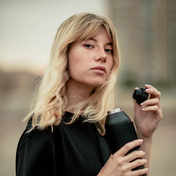 A person with shoulder-length blonde hair and freckles holds the Closca Wave Large - Black - 600ml flask, featuring curated Estonian design. Outdoors with a blurred background, they wear a black top, capturing modern Tallinn shopping vibes. Gate21.ee