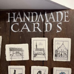 A wooden display labelled "Handmade Cards" in white showcases curated Labora - Postkaart cards, featuring hand-drawn Estonian designs of buildings and nature in black ink, arranged in two rows&mdash;ideal for a unique gift shop with a window behind. Gate21.ee