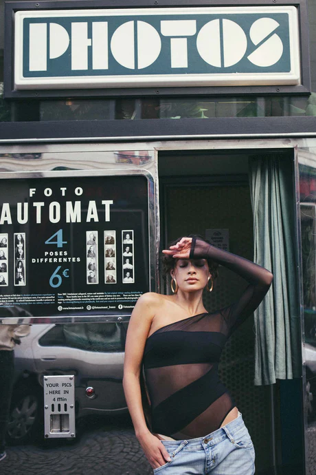 Wearing the Sex and the City Bodysuit and jeans, a person strikes a pose in front of a vintage PHOTOS booth—perhaps after browsing Tallinn’s unique shopping concept stores or finding gems at a local gift shop. Gate21.ee