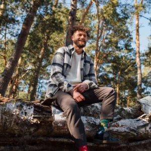 A man wearing Meet the Shrooms sits on a fallen tree amid tall forest trees and sunlight—his look inspired by curated, unique ideas. Gate21.ee
