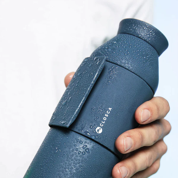 A hand holds the Closca Wave Large - Abyss - 600ml bottle with condensation. Its unique design and attached strap make it ideal for Tallinn shopping trips or concept store visits. A white shirt appears in the background. Gate21.ee