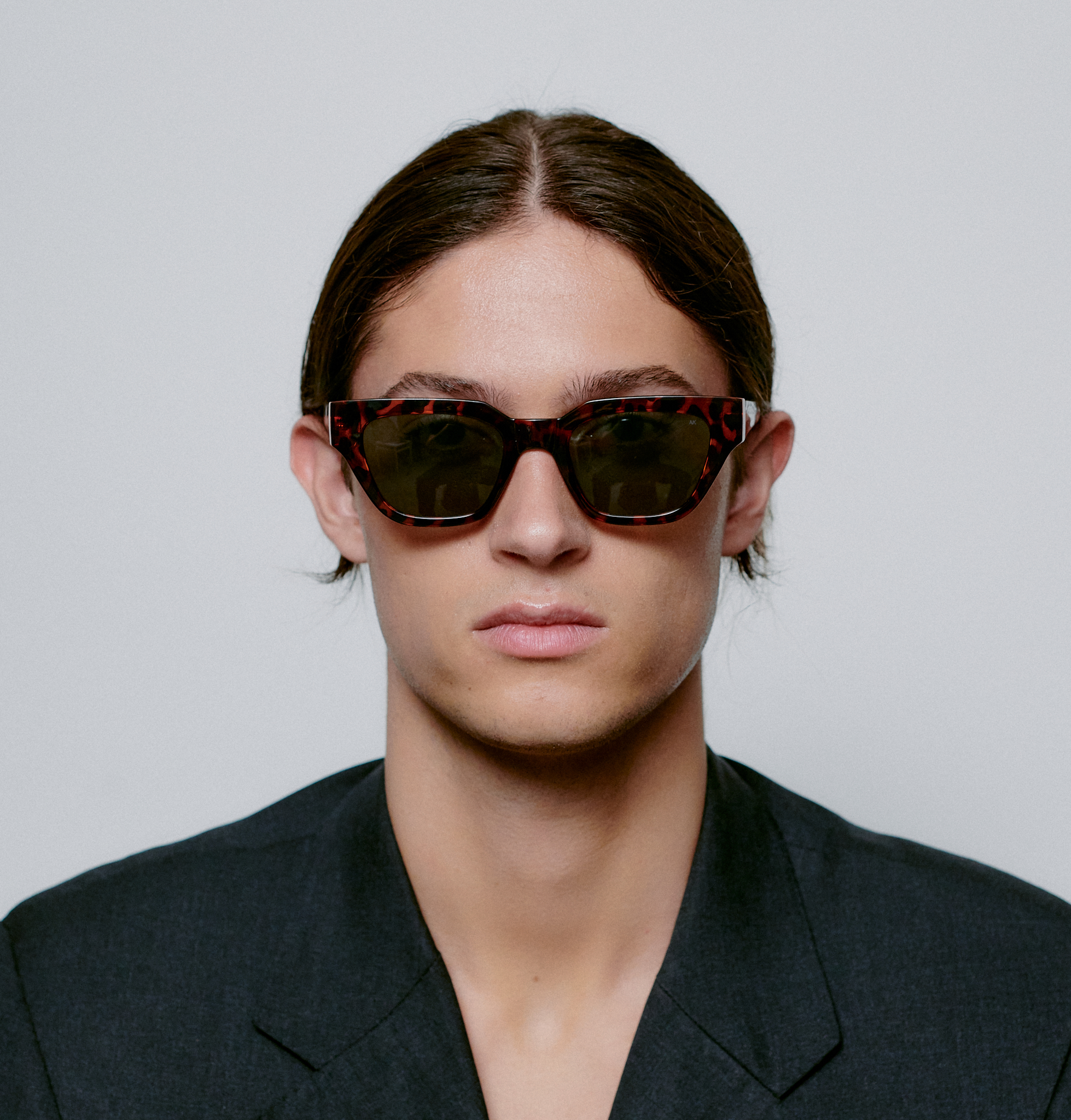 A person with straight brown hair parted in the middle, wearing KAWS - Demi Tortoise sunglasses and a dark blazer, embodies modern Estonian design whilst looking directly at the camera against a plain white background. Gate21.ee