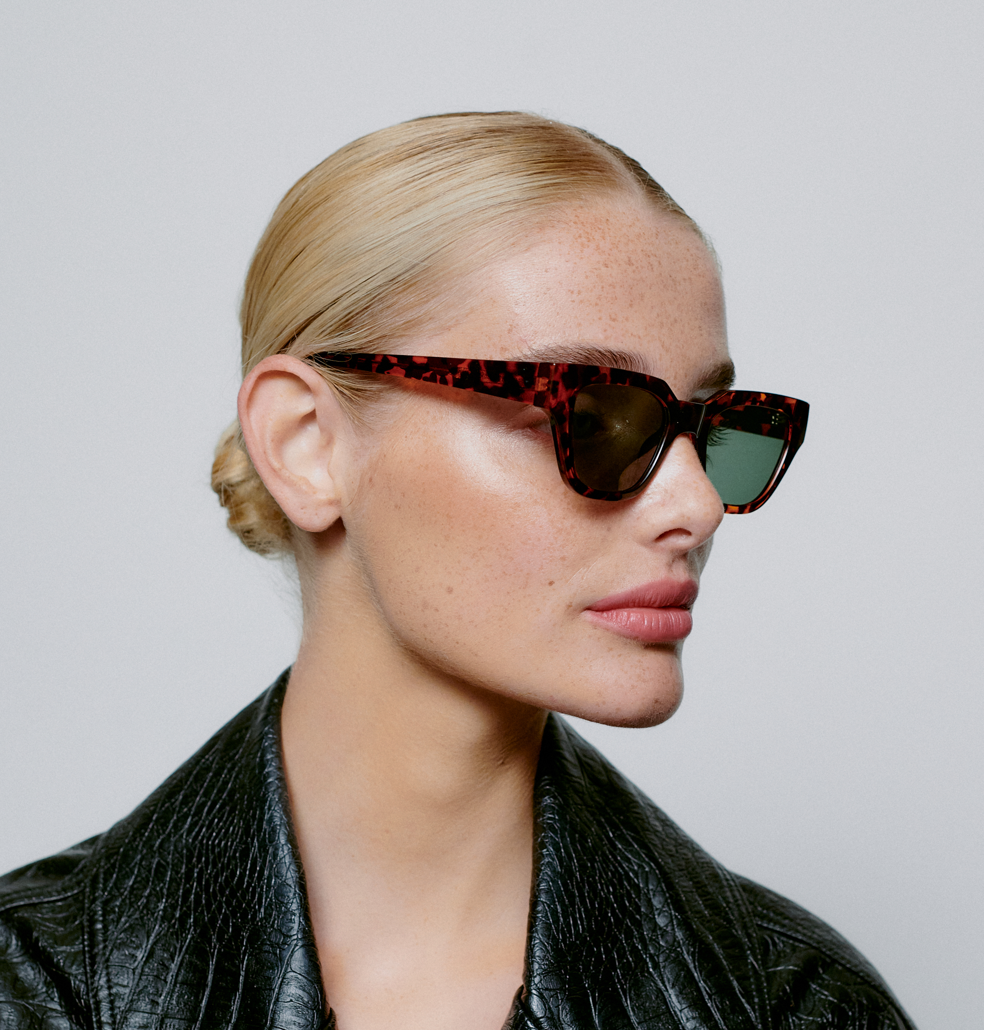 A person with slicked-back blonde hair and freckles wears KAWS - Demi Tortoiseshell sunglasses and a black textured jacket, shown in profile against a plain light background—highlighting the minimal aesthetic of Estonian design. Gate21.ee