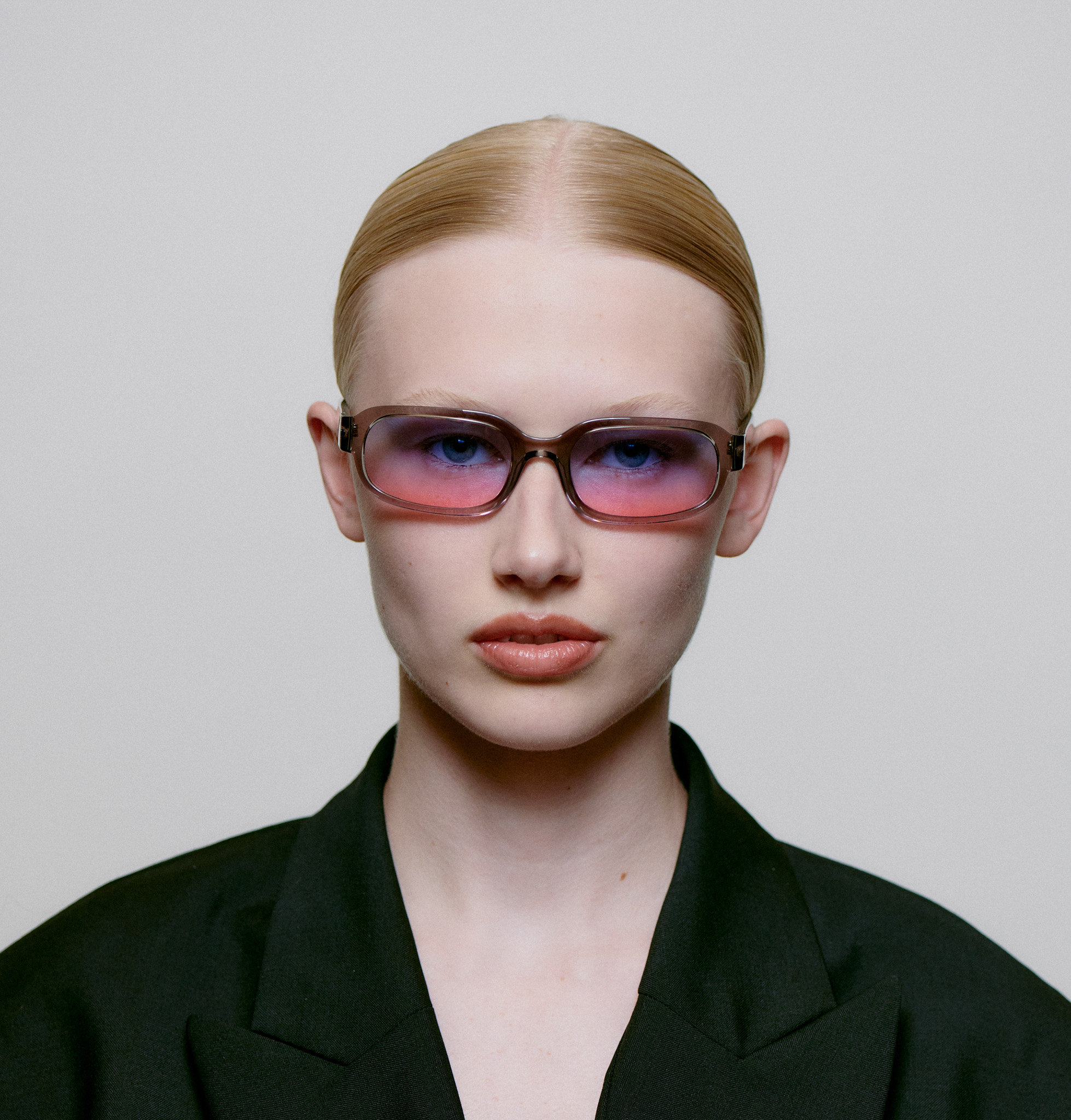 A person with slicked-back blond hair wears WILL - Grey Transparent sunglasses and a dark blazer, facing the camera against a plain light background—embodying the curated essence of modern Estonian design. Gate21.ee