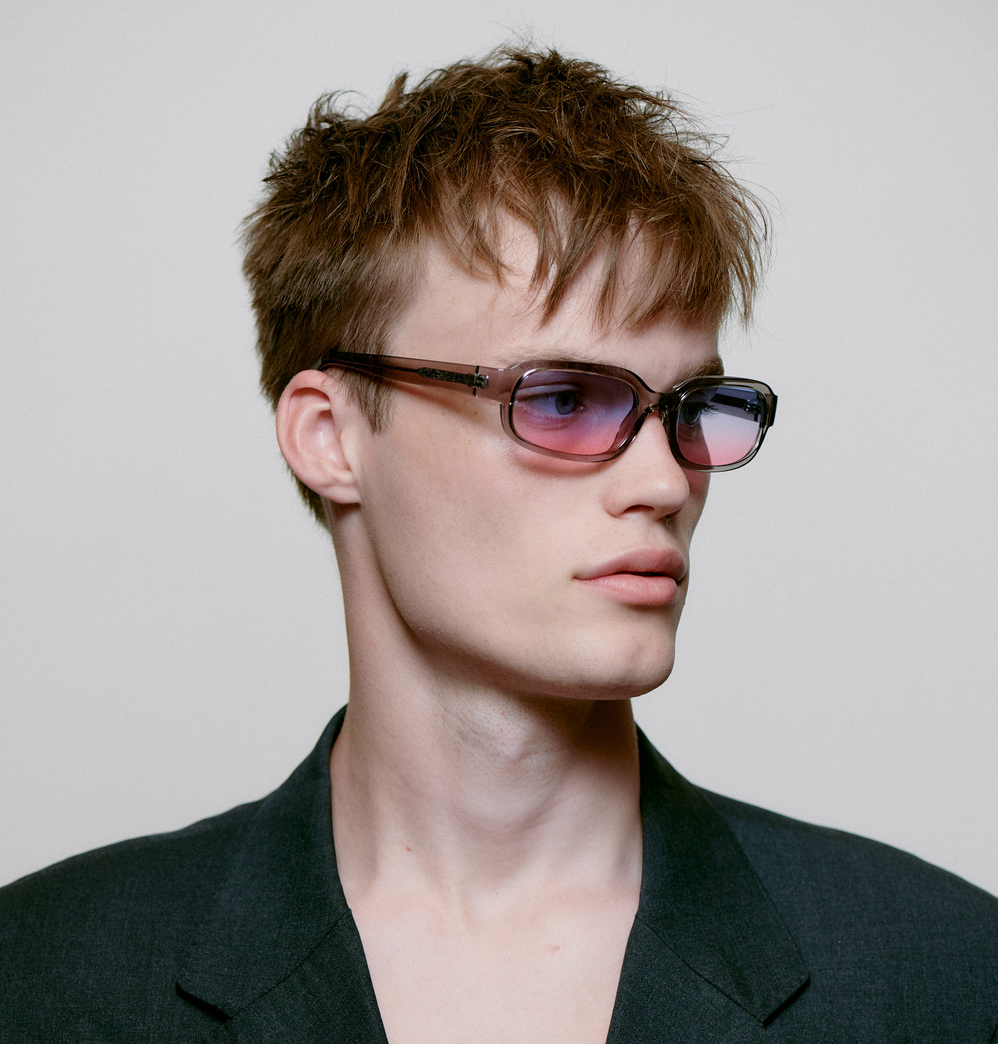 A person with short, tousled brown hair wears a dark blazer and WILL - Grey Transparent rectangular sunglasses, showcasing modern concept store style against a plain light background. Gate21.ee