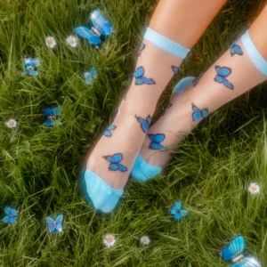 A person wears Butterfly - Transparent Socks featuring blue butterfly patterns and blue toe caps, standing on green grass with white daisies and blue butterfly decorations. Gate21.ee