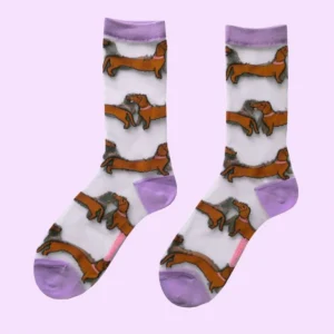 The Dachshund - Transparent Socks by Coucou Suzette are a pair of white see-through socks with lavender toes, heels, and cuffs, decorated all over with a repeating pattern of brown dachshund dogs featuring black ears and tails. The socks are shown laid flat on a light purple background. Gate21.ee