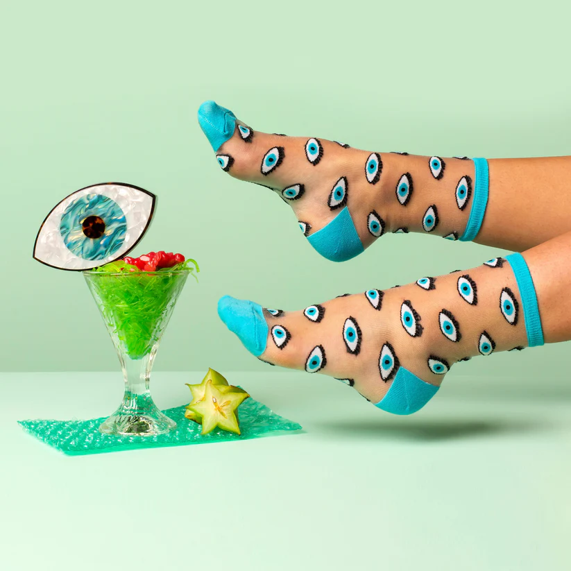 Blue Eyes - Transparent socks with blue eye patterns and turquoise trim are shown near a glass filled with green shredded material, a red object, and a paper eye decoration; a sliced carambola sits beside them—perfect for unique gift ideas. Gate21.ee