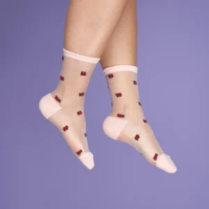 A pair of feet wears Ladybird - Transparent Socks featuring sheer fabric with light pink toe, heel, and cuff. Small red ladybirds decorate the socks, set against a solid purple background. An elegant Estonian design. Gate21.ee