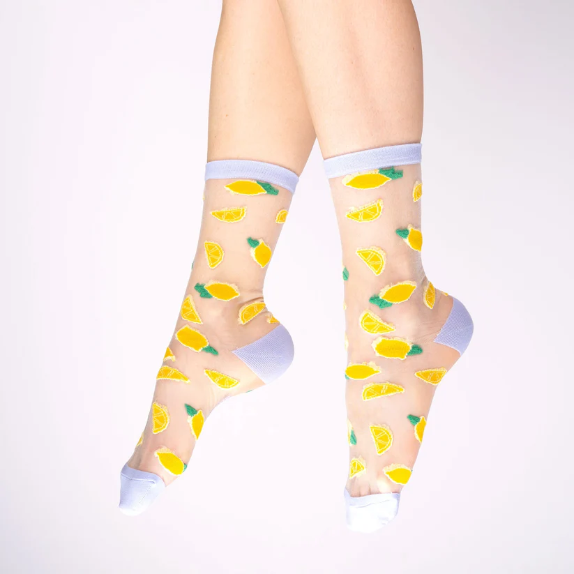 A pair of feet in Lemons - Transparent Socks, featuring yellow lemon wedges and green leaves, plus solid white toes, heels, and cuffs—a quirky gift or standout Tallinn shopping find on a simple light background. Gate21.ee