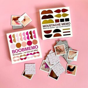 Two OMM - Boobmemo memory games on a pink surface—one features illustrated breasts, the other moustaches. Scattered cards add playful charm, offering curated Estonian design that's perfect for any concept shop. Gate21.ee