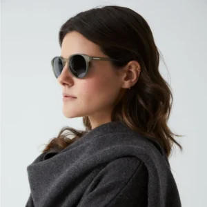 A woman with wavy brown hair wears round black "Isla" Moss Sunglasses and a dark grey coat, shown in profile against a plain light background, embodying the understated elegance of curated Estonian design. Gate21.ee