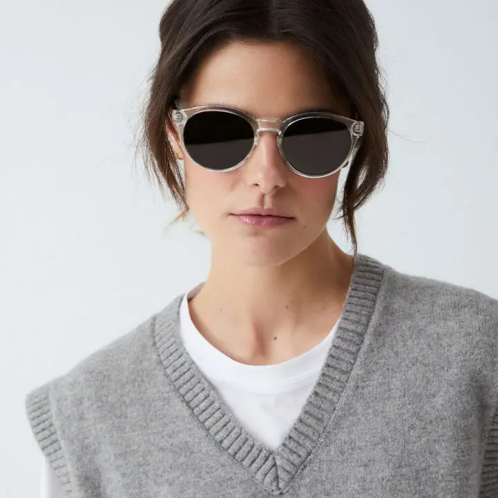A person with dark hair wears "Costa" Crystal Sunglasses, a white T-shirt, and a grey knitted waistcoat—a stylish look fit for a curated gift shop. The background is plain and light-coloured. Gate21.ee