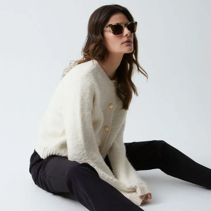 A woman with long brown hair sits on the floor against a plain backdrop, wearing "Colina" Havana Sunglasses, a cream fuzzy jumper with large buttons showcasing Estonian design, and black trousers. She gazes to the side, her posture relaxed. Gate21.ee