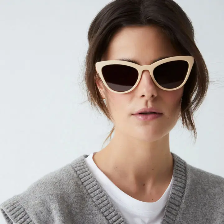 A person with dark brown hair sports the "Colina" Creamy Sunglasses, a white T-shirt, and a grey knitted jumper vest for an effortlessly curated look against a plain, light background. Gate21.ee