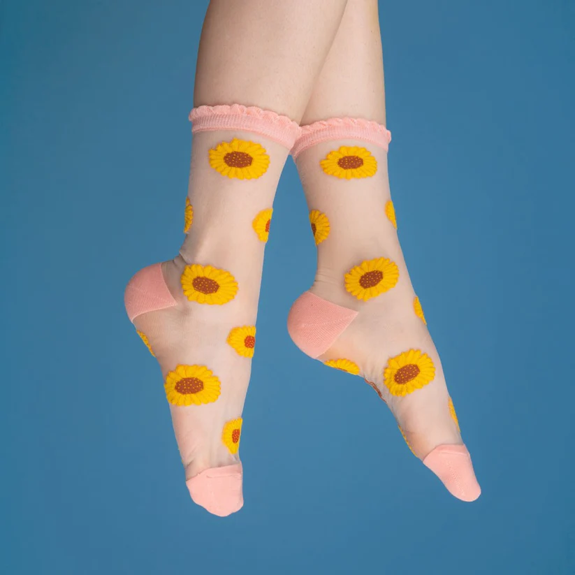 A pair of feet in Sunflowers - Transparent Socks, featuring yellow sunflower patterns and pink accents, posed against a blue background—highlighting curated Estonian design ideal for any gift shop. Gate21.ee