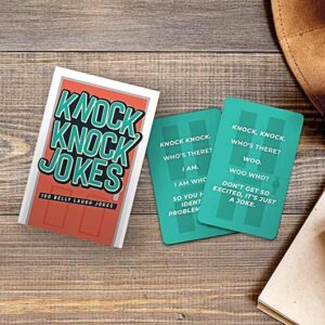 A box labelled "Knock Knock Jokes - 100 of the Best" rests on a wooden surface next to two cards displaying white knock-knock jokes, ideal for anyone after unique finds at a concept store. A brown object peeks into the corner. Gate21.ee