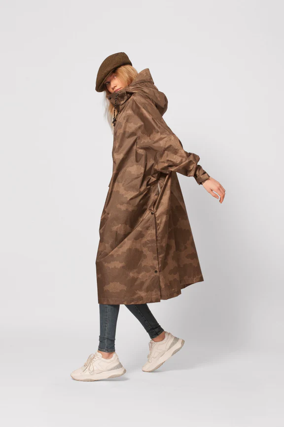 A person wearing The Clouds brown camouflage-pattern raincoat, grey skinny jeans, light trainers, and a brown flat cap poses sideways against a plain white background. Gate21.ee