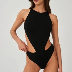 A woman models the Naked Instinct - Black, a black one-piece swimming costume with side cut-outs and a high neckline. Her long brown hair frames her neutral expression as she stands against a plain background, glancing to the side. Gate21.ee