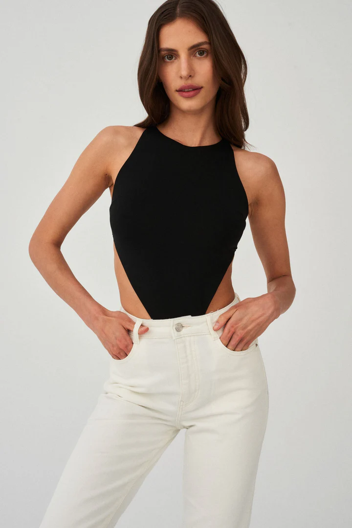 A woman models the Naked Instinct - Black sleeveless bodysuit with open sides, paired with high-waisted white jeans. Her long brown hair is loose and her hands are in her pockets, showcasing a chic Tallinn-inspired look against a plain background. Gate21.ee
