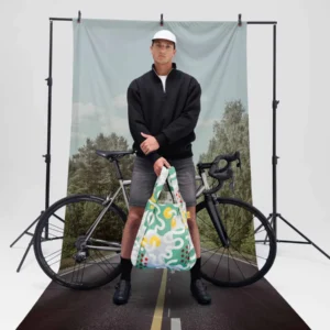 A man in a black jacket, shorts, and white cap stands in front of a bicycle, holding the Tour de France - Special Edition bag. A nature backdrop and road graphic complete the scene, evoking a cycling adventure. Gate21.ee