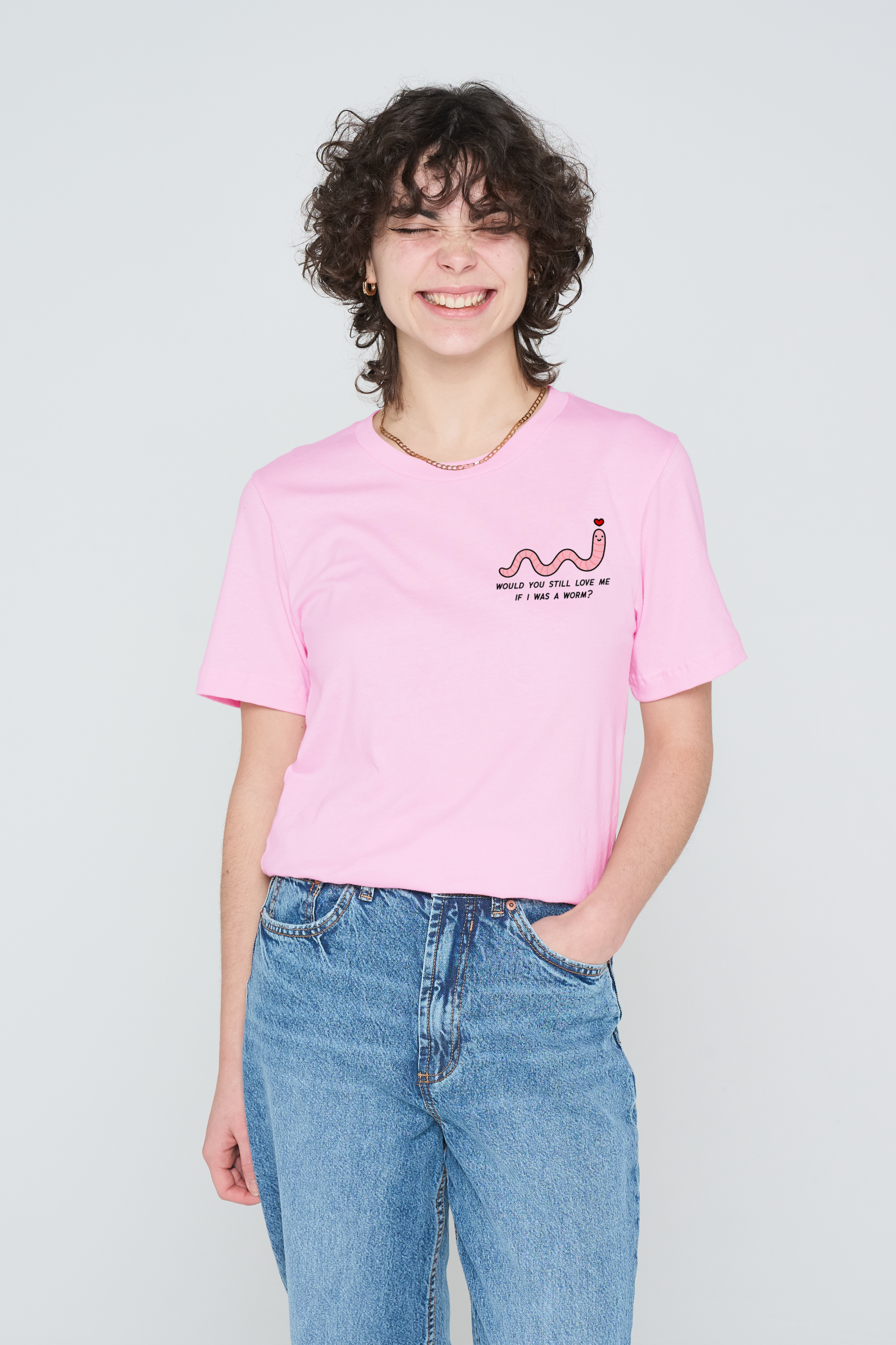 A person with short curly hair smiles with eyes closed, wearing the pink Love Me Worm T-Shirt featuring a small worm graphic and text, paired with blue jeans, standing against a plain light background. Gate21.ee