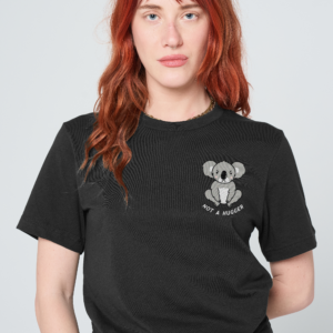 A person with long red hair stands against a plain background, wearing a pink Love Me Worm T-Shirt featuring a cheerful cartoon worm and the phrase Love Me. Reminiscent of the playful Sassy Spud style, they have a neutral expression and are wearing blue jeans. Gate21.ee