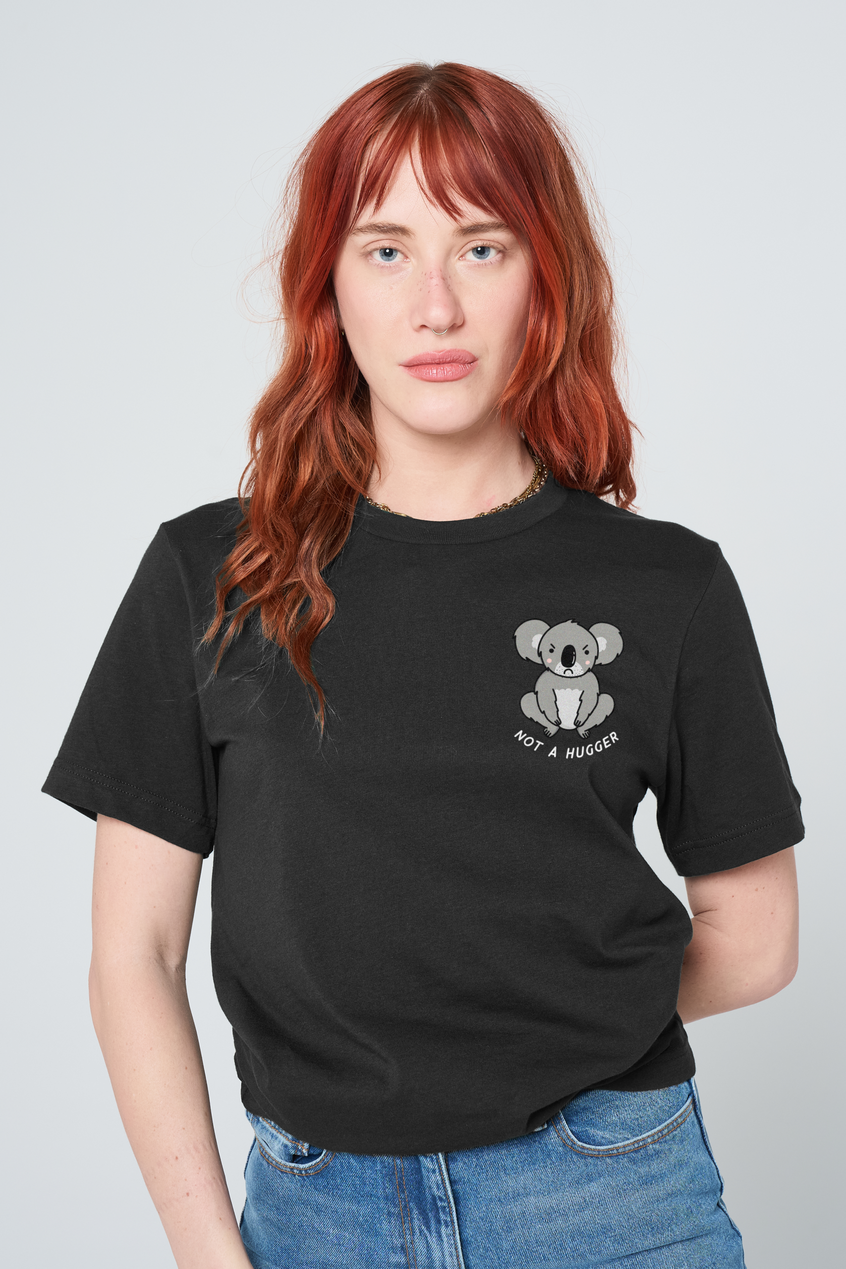 A person with long red hair stands against a plain background, wearing a pink Love Me Worm T-Shirt featuring a cheerful cartoon worm and the phrase Love Me. Reminiscent of the playful Sassy Spud style, they have a neutral expression and are wearing blue jeans. Gate21.ee