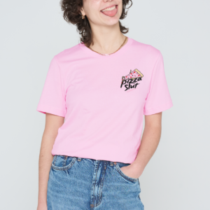 A person with curly hair, wearing a sassy pink Love Me Worm T-Shirt and blue jeans, stands against a plain white background, smiling with their tongue out and one hand in their pocket, radiating the same confident vibe as you’d find in the Love Me Worm T-Shirt – Pink. Gate21.ee