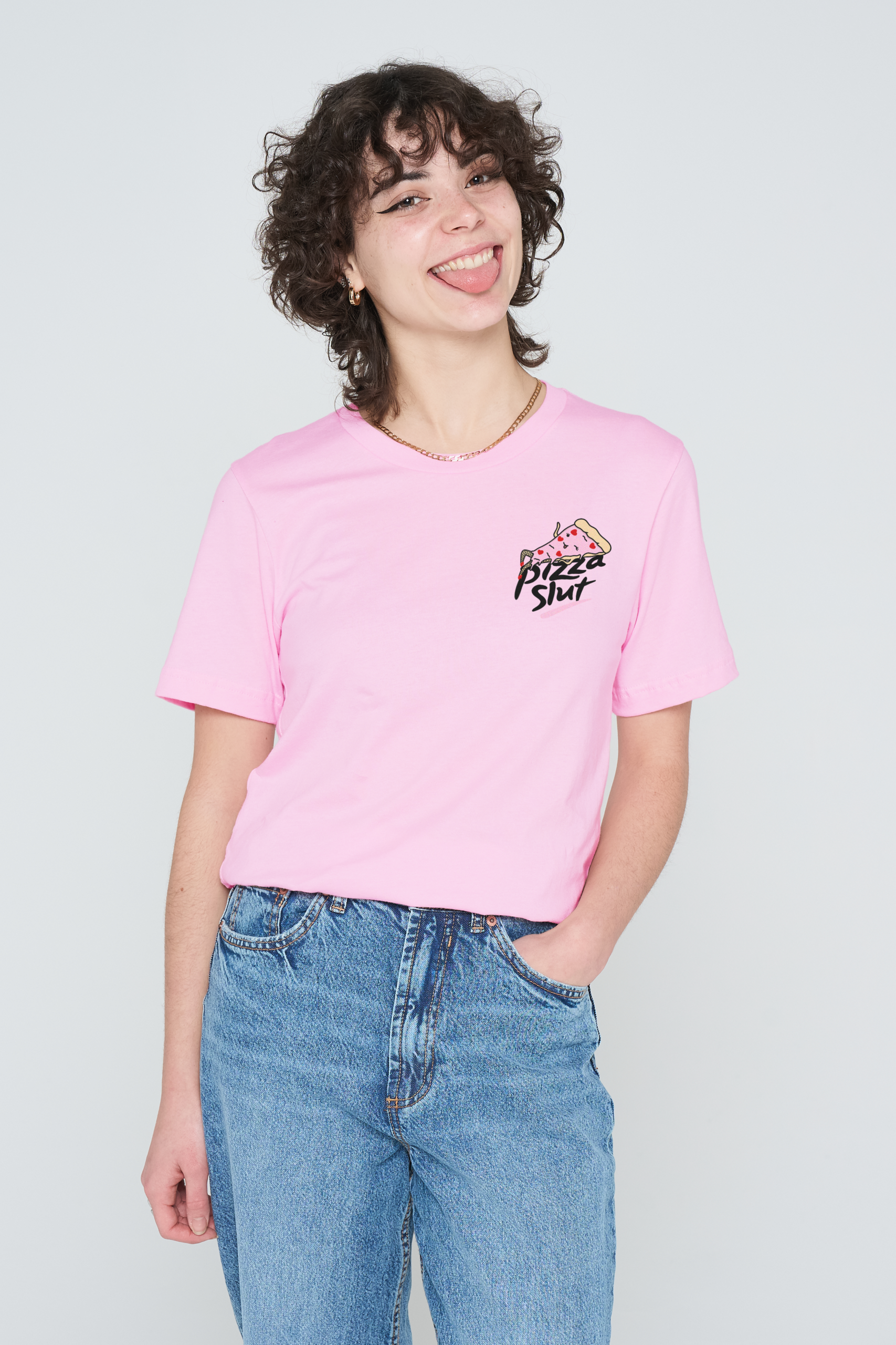 A person with curly hair, wearing a sassy pink Love Me Worm T-Shirt and blue jeans, stands against a plain white background, smiling with their tongue out and one hand in their pocket, radiating the same confident vibe as you’d find in the Love Me Worm T-Shirt – Pink. Gate21.ee