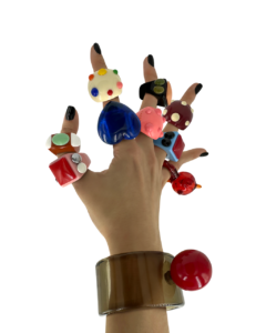 A hand with black-painted nails wears the colourful, chunky Spring Dragon Fruit - Ring and a large translucent brown bracelet with a red sphere, set against white—a bold Estonian-inspired look perfect for Tallinn or your favourite concept store. Gate21.ee