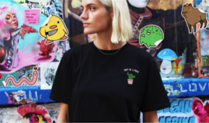 A person with short blonde hair wears the Bananciaga Embroidered T-Shirt in black, featuring a small potted plant and "DON'T BE A PRICK" text, standing before a vibrant graffiti wall&mdash;ideal for Tallinn shopping inspiration. Gate21.ee