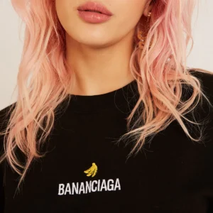 A person with wavy, pastel pink hair wears the Bananciaga Embroidered T-Shirt in black, featuring a yellow banana and "BANANCIAGA" in white—just the lower face and upper torso are shown, evoking chic Tallinn boutique style. Gate21.ee